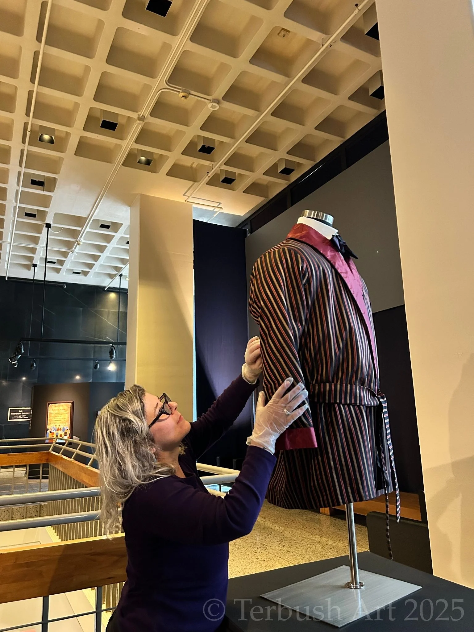 A modern hunting jacket colored with black, gold, and red vertical stripes is on a raised half-mannequin. A belt with the same pattern is tied around the waist of the jacket on the display. Kim Terbush is inspecting the elbow of the jacket to ensure there is no wear or damage to the item.